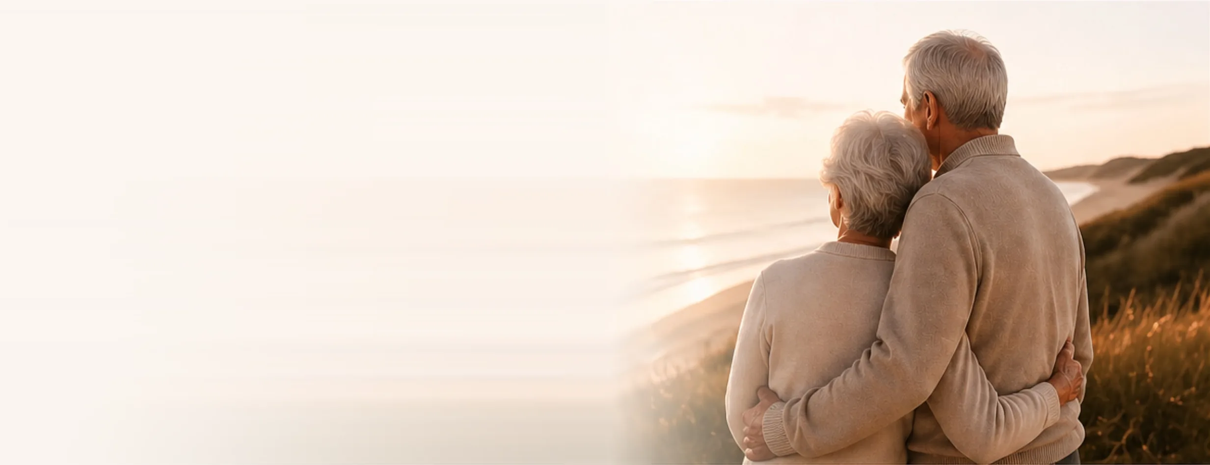 Ein älteres Ehepaar steht Arm in Arm in den Dünen und blickt bei Abendlicht auf das Meer