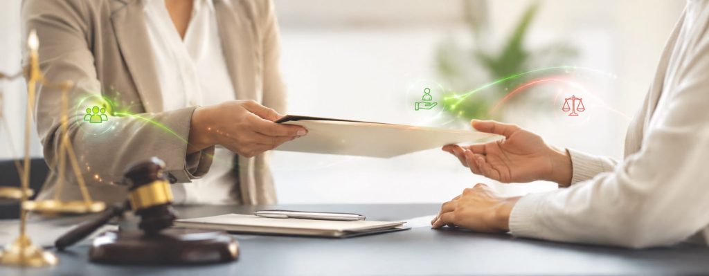 Beraterin reicht einer Mandantin eine Mappe über den Schreibtisch, daneben Waage und Richterhammer